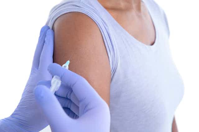 A woman being injected in her arm by a pair of hands in purple gloves.