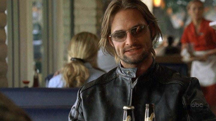 Sawyer (Josh Holland) sits in a restaurant booth, wearning aviator sunglasses and a leader jacket zipped up to his neck.