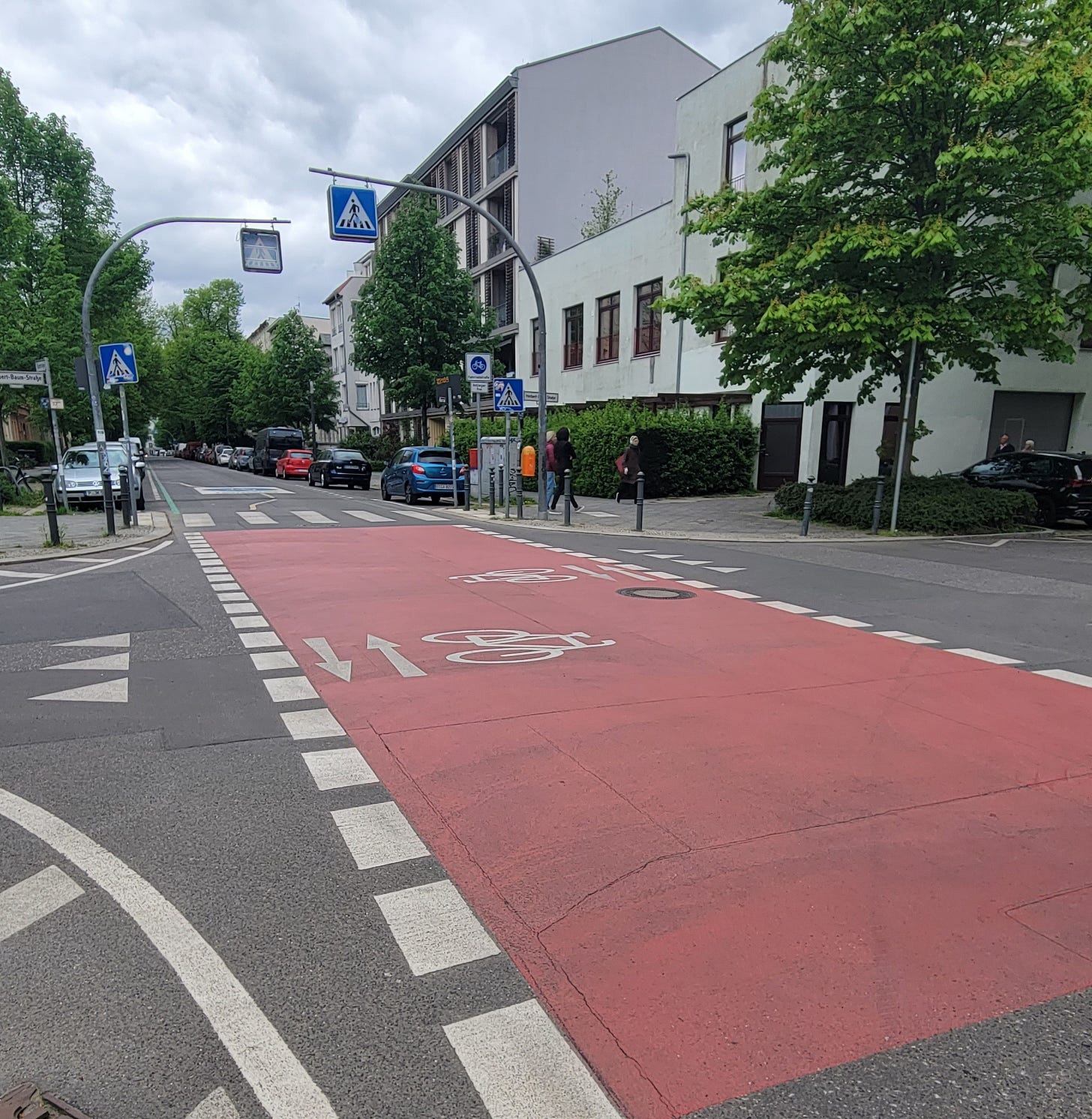 Street with painted markings signifying it is for use by bikes only. The bike street has red-painted pavement with white highlights marking the direction of travel. Blue signs hanging over the street warn of a pedestrian crossing ahead.