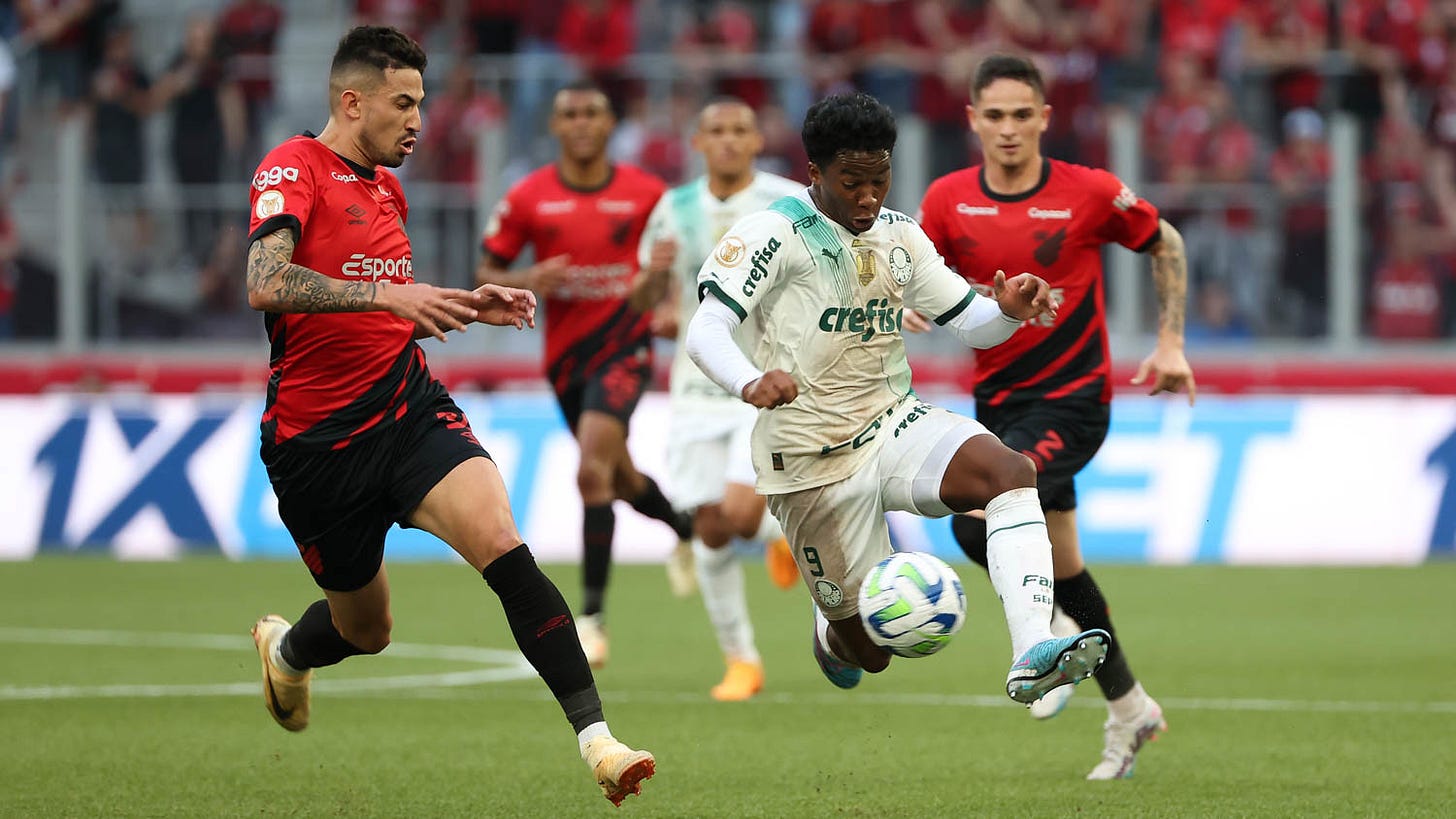 Imagem mostra Endrick com a bola, perseguido por um jogador do Athlético. Endrick veste uniforme do Palmeiras todo branco, com detalhes em verde, e o jogador do Athlético, à esquerda dele, usa camisa vermelha com detalhes em preto, calções e meias pretas. Os dois estão de frente. Ao fundo, desfocados, aparecem mais um jogador do Palmeiras e dois do Athlético.