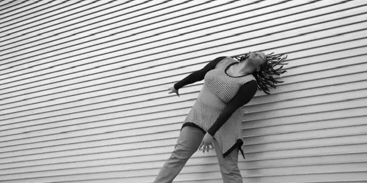 A black & white dance image of Kayla, who is a dark-skinned black woman. She is in front of a textured wall that has horizontal layers. Her arms are energetically reaching down to the diagonal as her head is tilting to the diagonal with her dreads moving back in that same direction. She is wearing a diagonally striped long sleeve shirt with pants.