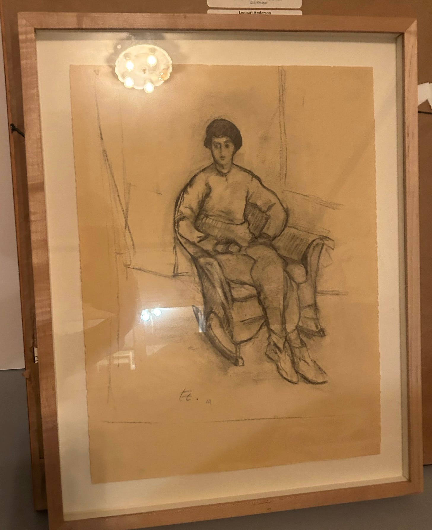 A framed drawing of a woman seated with legs crossed in a wicker rocking chai, framed, the glass reflecting light fixtures in the room. 