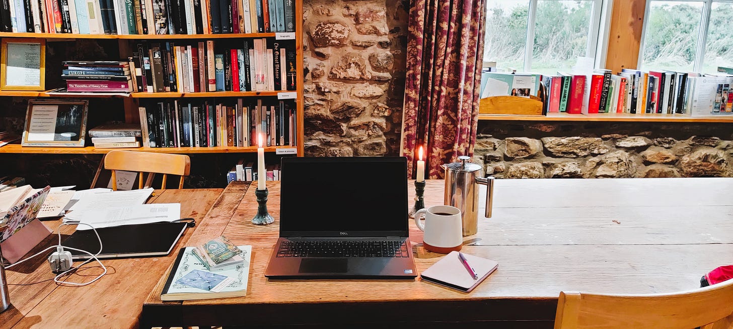 A long wooden dining table with a laptop, two candles, coffee and writing implements on top, To the left, someone else's messy work station. A book case behind the table and a window on the right