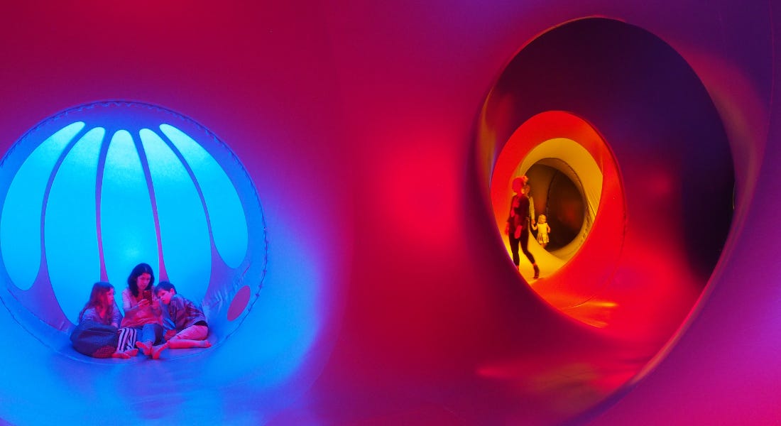 A purple and red luminarium space with people sat in the blue window and people exploring it