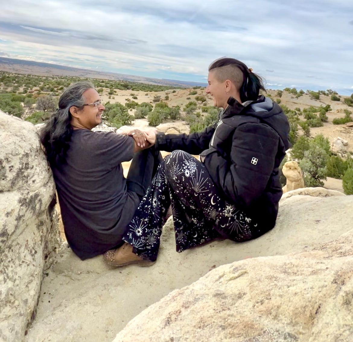 Lyric and David, sitting on top of the sand stone rocks in the New Mexico Desert. They both are dressed in black, looking at each other, smiling. 