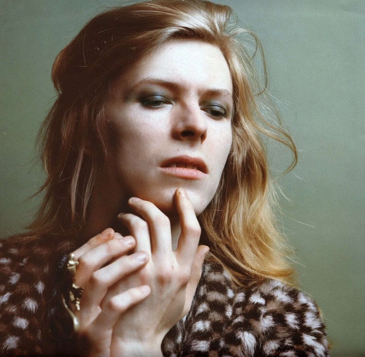 David Bowie with shoulder-length blond hair, wearing eyeshadow, rings, and a dyed brown and white fur coat
