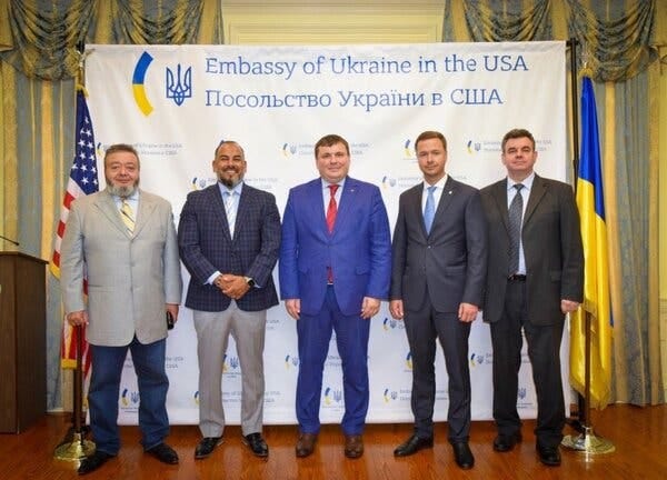 Five men in suits stand in front of a backdrop reading, “Embassy of Ukraine in the USA.”