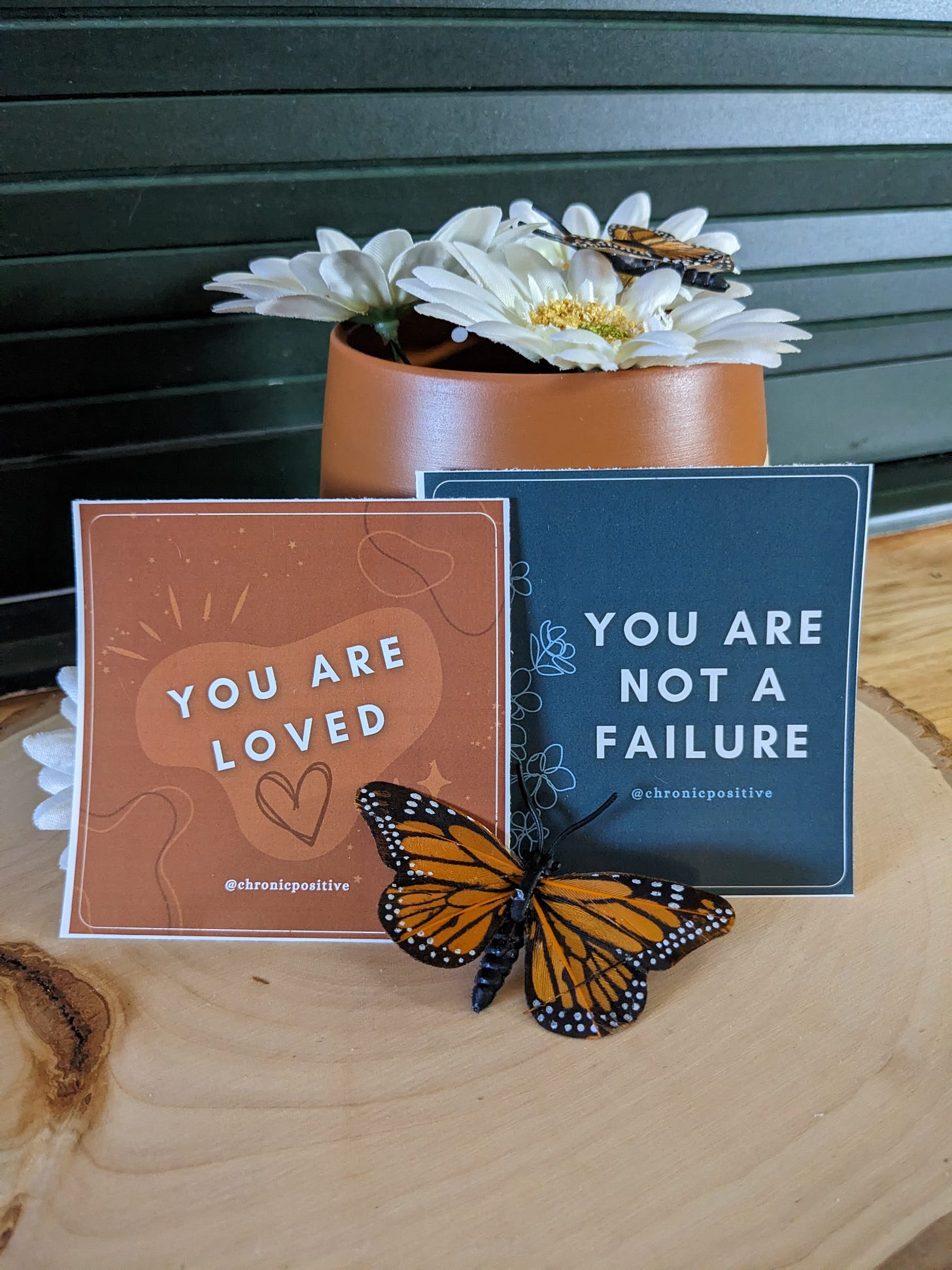 A photo of an orange sticker that says "you are loved" on a background accented with blobs and swirls and a blue sticker that says "you are not a failure" with an artsy floral line drawing. The stickers are staged with an earthy orange vase, daisies, and fake monarch butterflies.