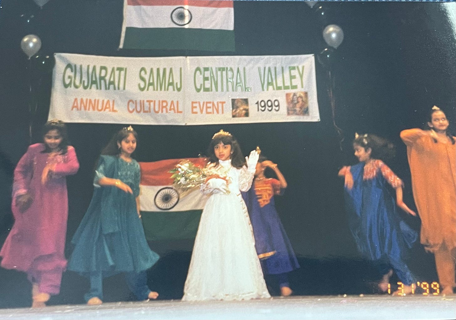 A 5-year-old Indian girl wears a white dress, tiara, and sash as she stands onstage. Surrounding her are other Indian girls, dancing in salwaar kameez outfits of differing colors. 
