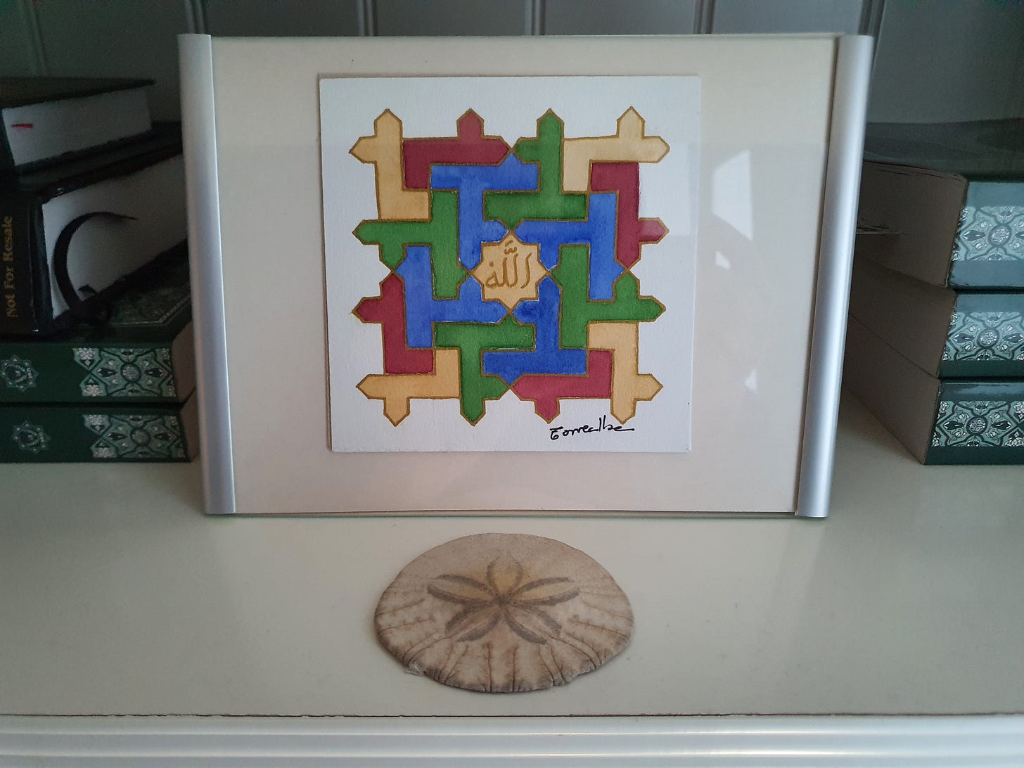 Framed image of interlocking multicolored angled lines surrounding the Name of Allah SWT in Arabic. In front, a sand dollar. In the background, books stacked horizontally. 