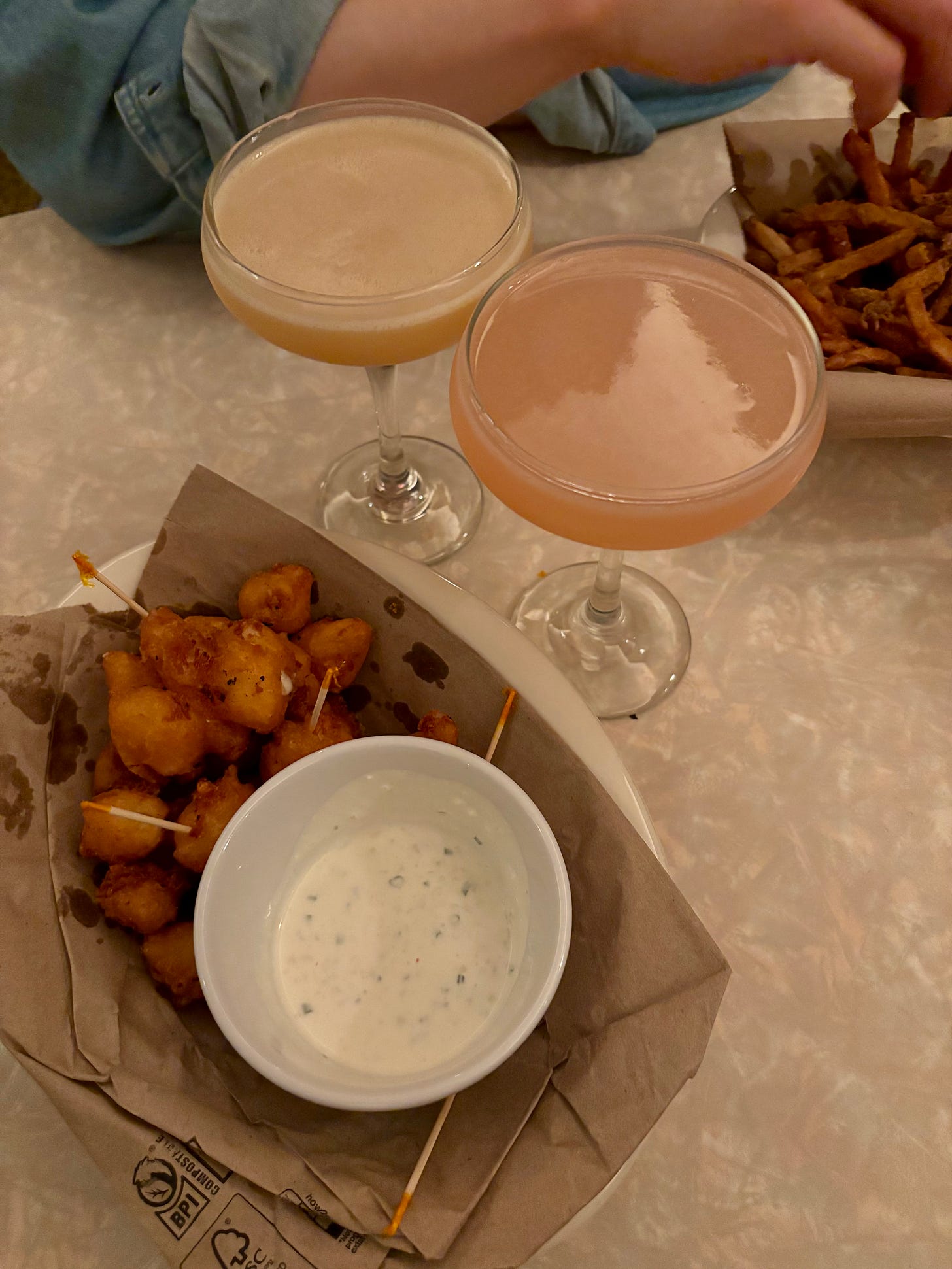 A basket of cheese curds and two coupe glasses containing brown and pink hued cocktails respectively. In the upper left, a hand reaches for french fries. A basket of cheese curds and two coupe glasses containing brown and pink hued cocktails respectively. In the upper left, a hand reaches for french fries.