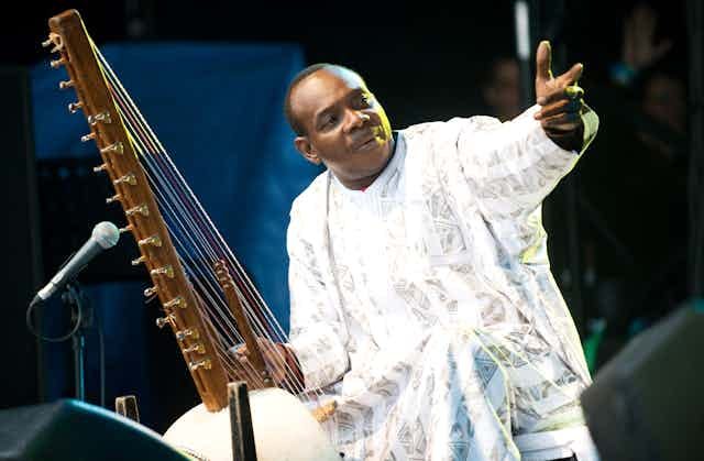 A man in an ornate white robe holds a hand up as he sits at a microphone with a large stringed musical instrument, apparently on a stage.