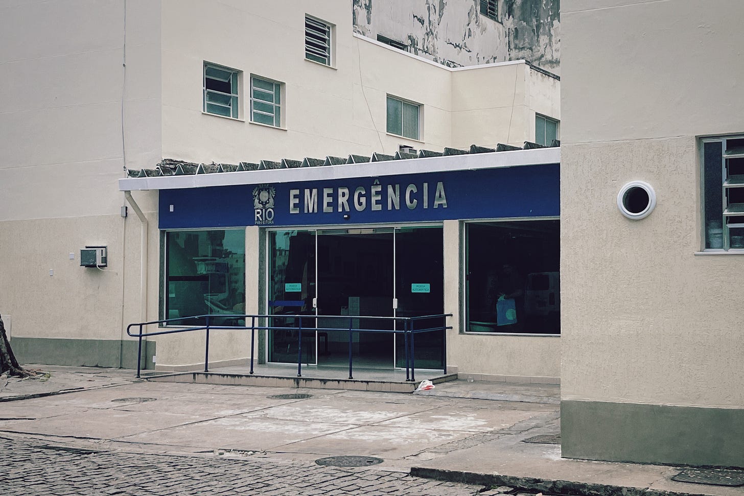 Photograph of a hospital on the outskirts of Rio de Janeiro. The building is white. Over a glass door is a blue sign that says EMERGENCIA in silver lettering, next to Rio's city seal.