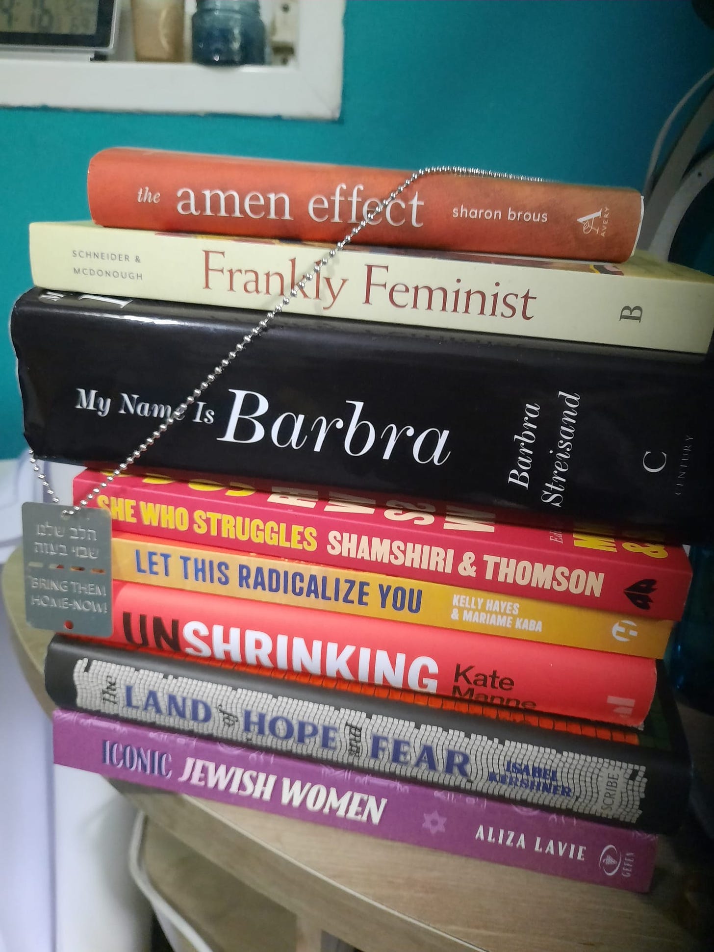 A pile of books on a desk, from top to bottom: The Amen Effect, My Name is Barbra, She Who Struggles, Let This Radicalize You, the Unshrinking, The Land of Hope and Fear, Iconic Jewish Women. With a chain/dogtag hanging around it, the chain of hostages.