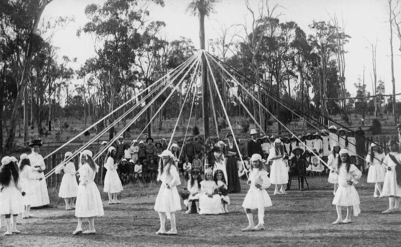 Children maypole dancing, 1900-1910 Children's group maypole dancing, 1900-1910. Girls stand in a circle around the pole and hold the ribbon streamers. They wear headgear, dresses, stockings and shoes. Two of the three girls sitting at the centre of the pole have flowers in their hair and hold baskets of flowers. A wreath hangs from the pole above their heads. A man sits and play the accordion to the right. Children are lined up against the fence and look on. Trees are visible in the background.