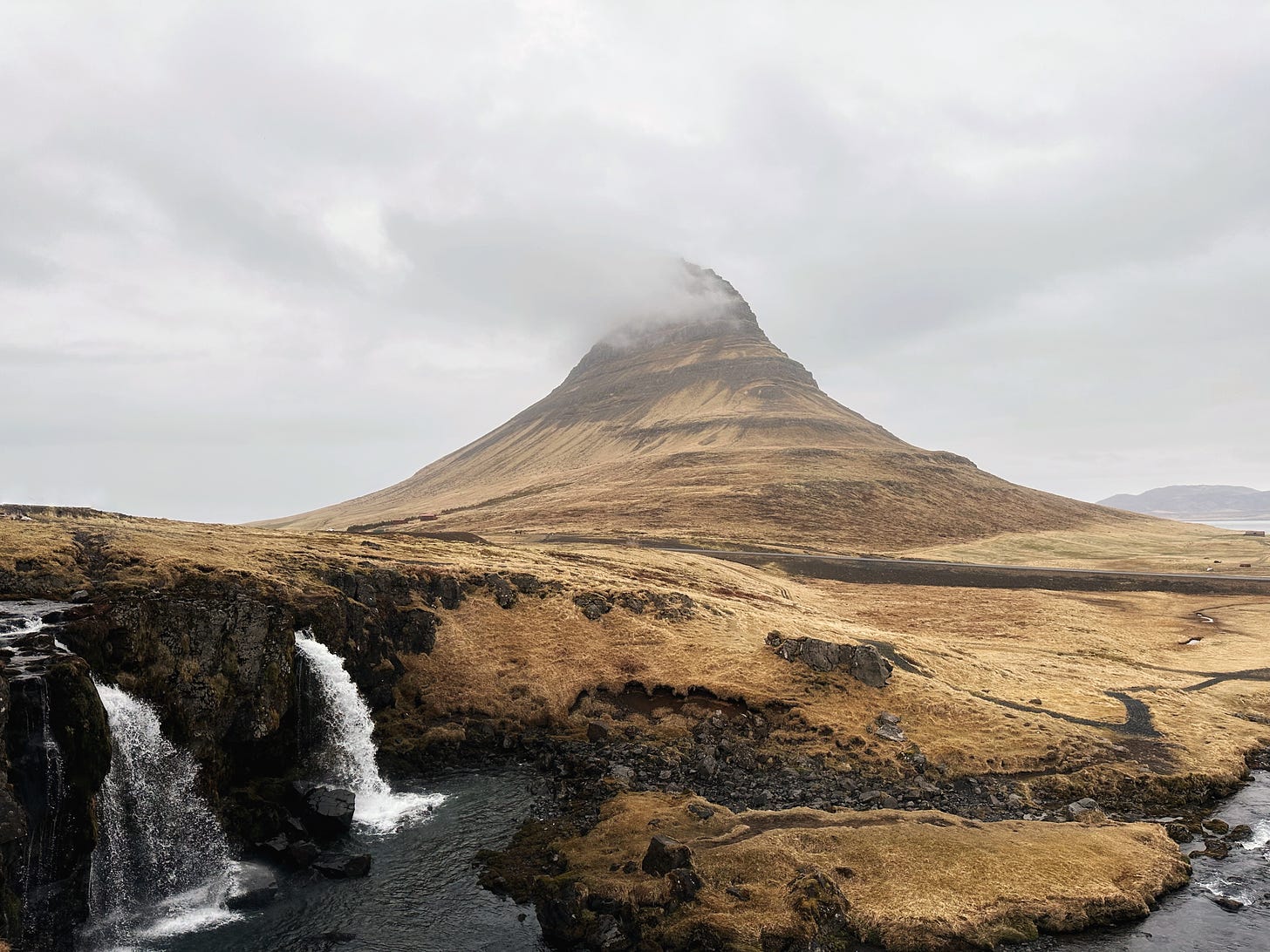 Kirkjufell mountain and Kirkjufellsfossar waterfalls.  The wedge shaped mountain is in the background covered in golden brown grass with wispy white clouds floating across the pointy tip. The waterfalls flow on the left side down into deep blue black water running below across the front of the image and out to sea across the right side of the frame. The sky is filled with multi colored gray clouds.