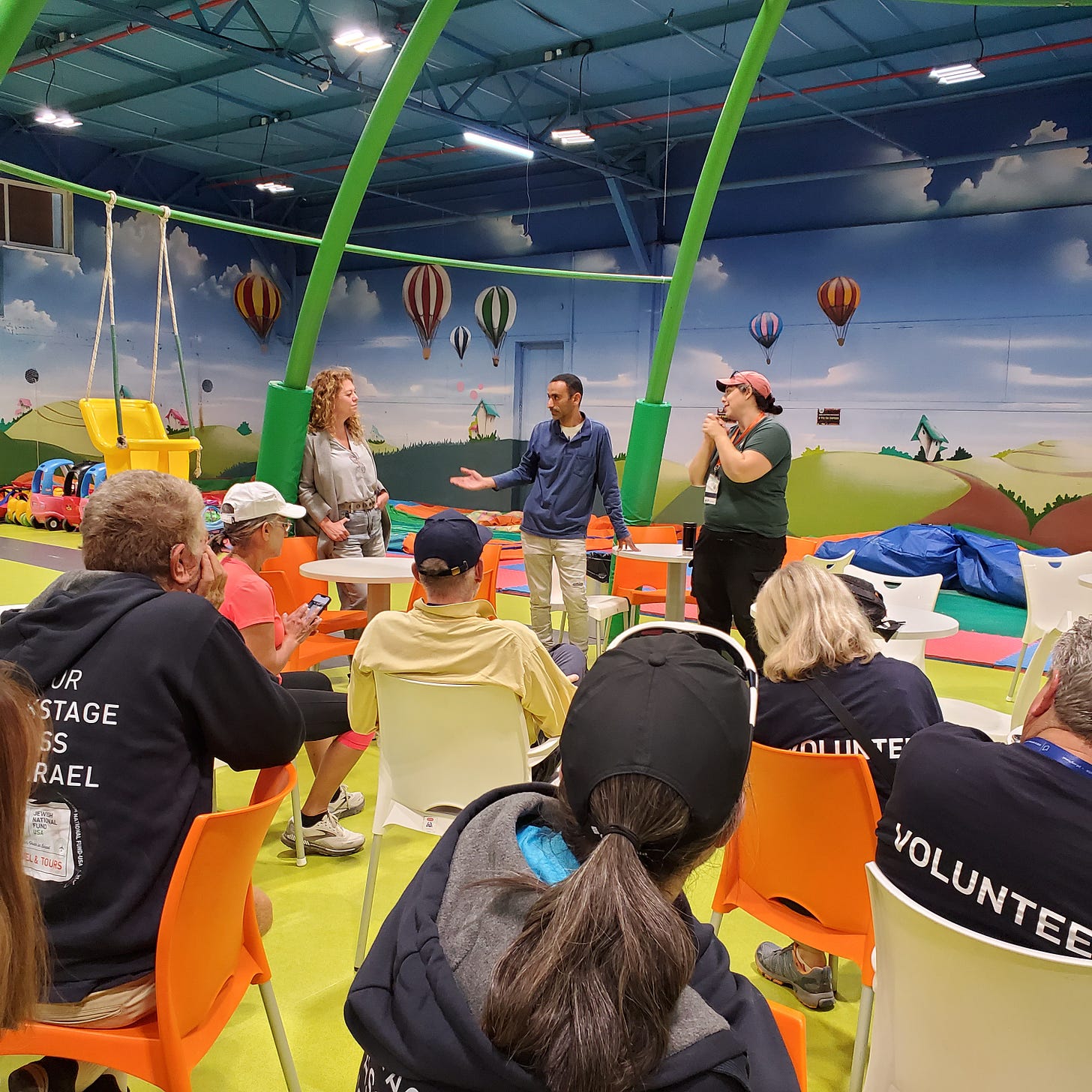 A seated group listens to three speakers describe the purpose and history of the indoor playground surrounded by colorful murals. 