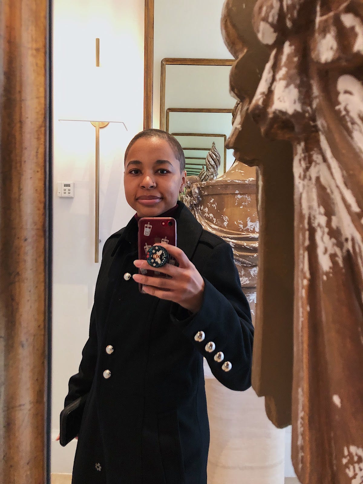 Girl taking a selfie in a large mirror with a huge statue of a woman in front of it. The girl has a shaved head, and is in a nice black pea coat.