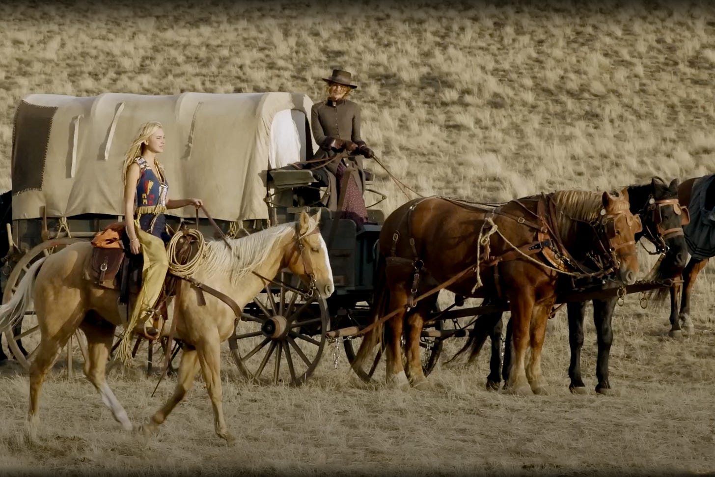 Elsa Dutton (played by Isabel May) rides a light-colored Palomino horse in traditional clothing through a dry grass landscape, while in the background her mother Margaret Dutton (played by Faith Hill) sits on the driver's seat of a covered wagon pulled by dark draft horses – a scene from the Western series “1883.” Elsa Dutton (played by Isabel May) rides a light-colored Palomino horse in traditional clothing through a dry grass landscape, while in the background her mother Margaret Dutton (played by Faith Hill) sits on the driver's seat of a covered wagon pulled by dark draft horses – a scene from the Western series “1883.”