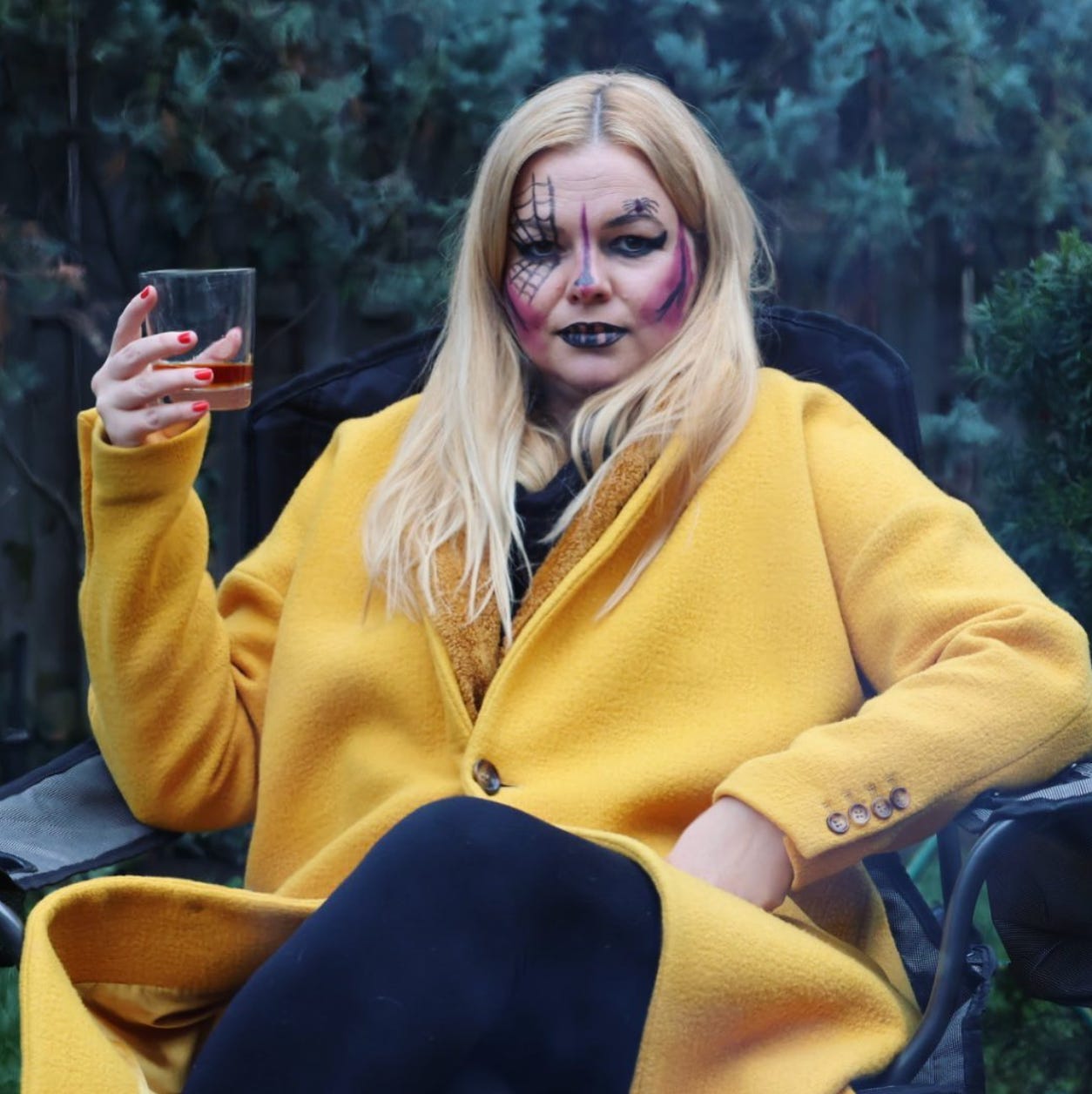 Amber sits in a chair outside. She wears a bright yellow coat and holds a glass of bourbon. She wears black lipstick and also has dark shades of lipstick used like face paint all over her face.