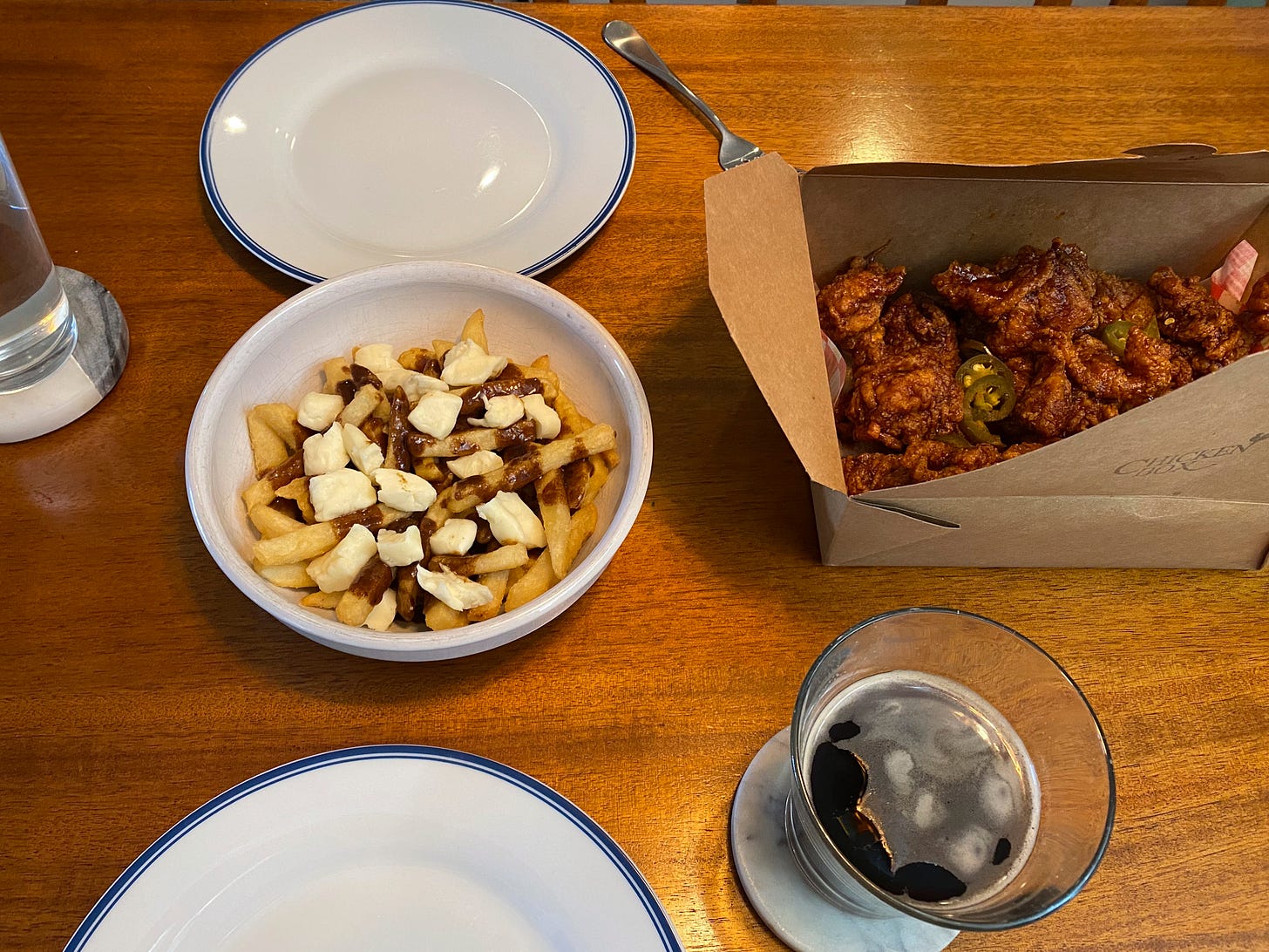 Two plates across from each other on the table. Between them is a white bowl filled with poutine, and a paper takeout box of chicken pieces in a spicy sauce, with pickled jalapeños sprinkled over top.