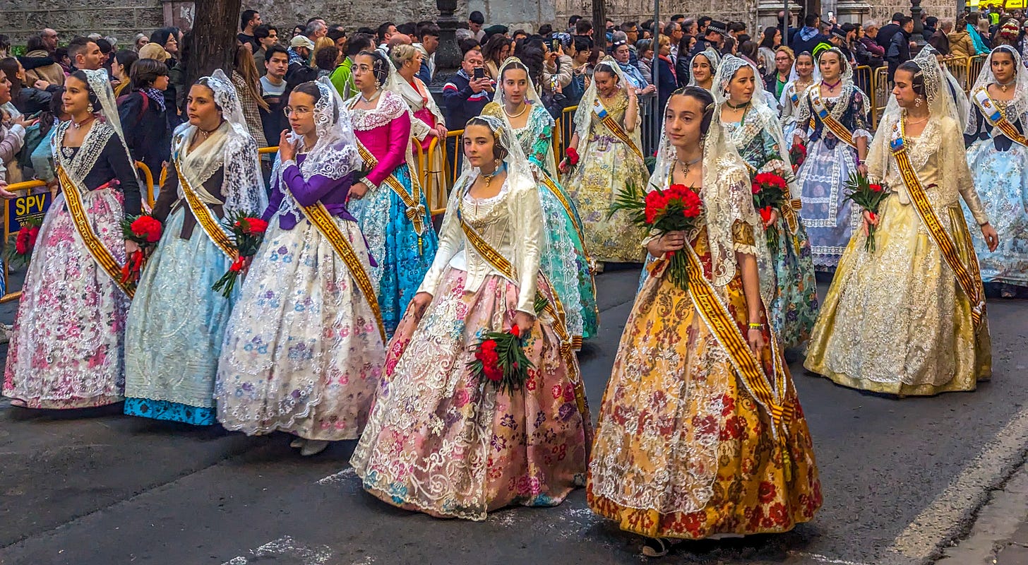 Young women wearing brightly colored dressses, white headdresses, and carrying bouquets of flower marching down the street. 