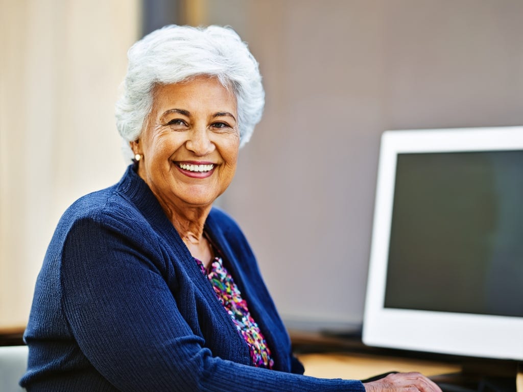 Portrait of smiling white-haired woman in blue sweater at her desk with comper screen to her left in background.