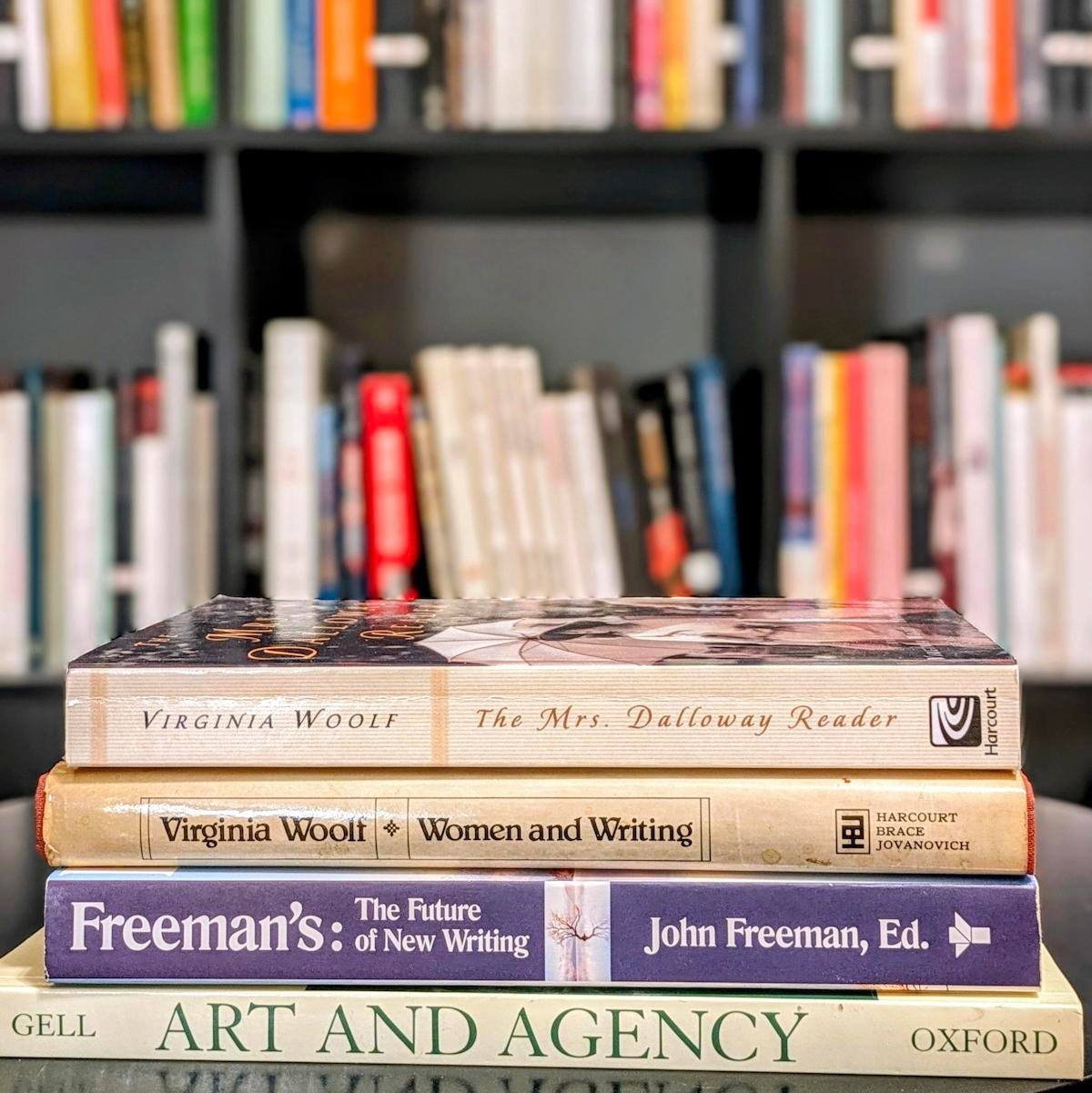 A pile of four books in front of a blurred out bookshelf. The books on the pile are: Virginia Woolf, The Mrs. Dalloway Reader; Virginia Woolf, Women and Writing; Freeman's: The Future of New Writing; Gell, Art and Agency.