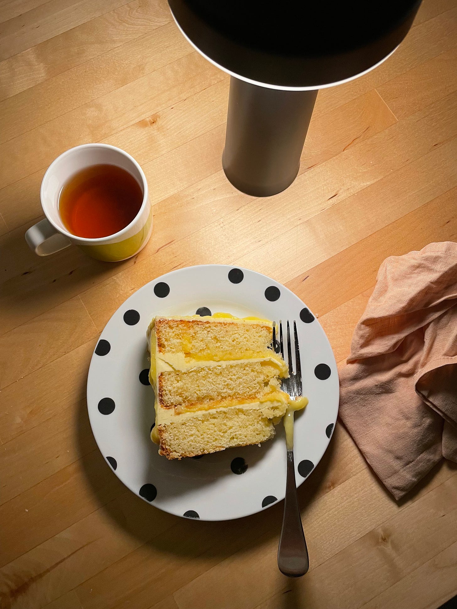 Slice of vanilla and lemon curd layer cake on a spotted plate. A small peppermint tea and lamp on a dining table accompany it.