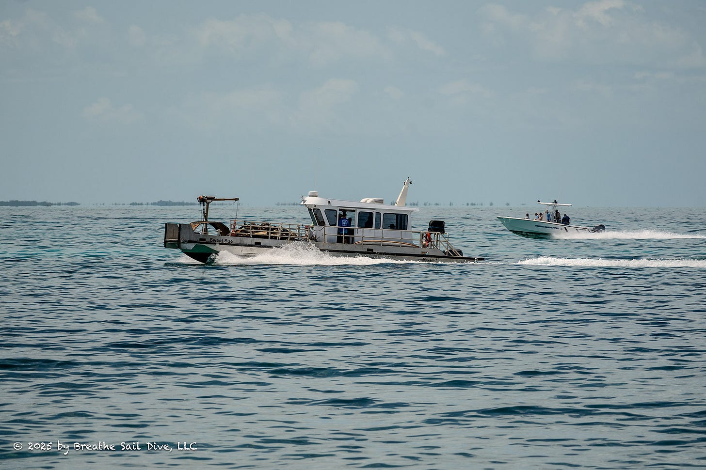 Two boats speed across calm, clear waters near Fowl Cays National Park. In the foreground, a workboat labeled “Sweet Sue” carries equipment and volunteers engaged in the wreck removal effort. A smaller powerboat with additional crew follows close behind. The scene reflects active grassroots efforts to address the stranded barge and tugboat in a remote marine environment. Two boats speed across calm, clear waters near Fowl Cays National Park. In the foreground, a workboat labeled “Sweet Sue” carries equipment and volunteers engaged in the wreck removal effort. A smaller powerboat with additional crew follows close behind. The scene reflects active grassroots efforts to address the stranded barge and tugboat in a remote marine environment.