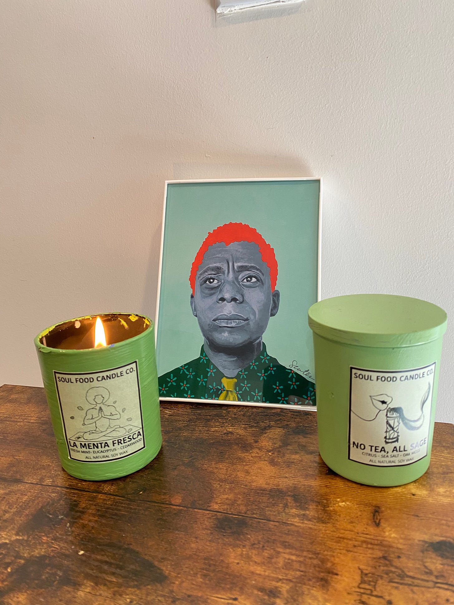 On a brown wood surface and against a white wall are two green candle jars. One has a shut lid and the other is open and aflame. Between them is an art deco artwork of James Baldwin.