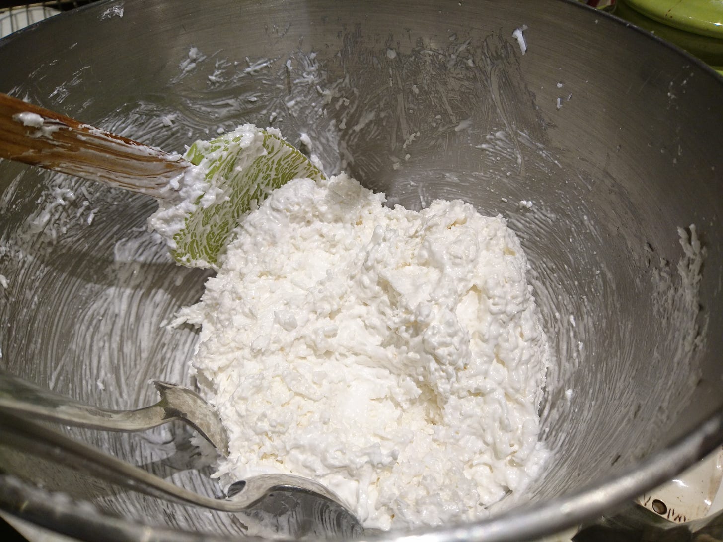 Egg white with bits of coconut sit in the bottom of a stainless steel bowl with two spoons and a silcon spatula