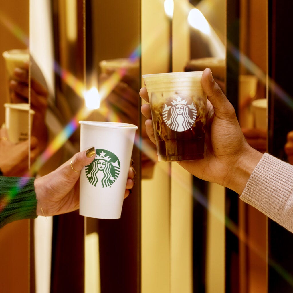 A hot Starbucks cup and a cold Starbucks cup with coffee held in front of shiny gold glass doors.