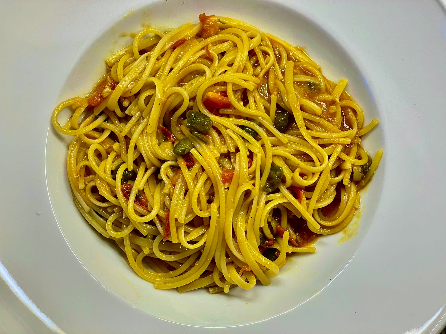 A plate of spaghetti with a rich tomato-based sauce, capers, anchovies, garlic, and chili, topped with fresh baby tomatoes and a drizzle of olive oil, served on an outdoor balcony.