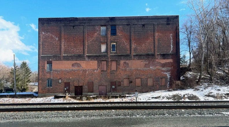 Entrep$ot agricole américain du XIX° siècle, façade en brique rouge, devant une route goudronnée sur laquelle passe une voie ferrée désaffectée