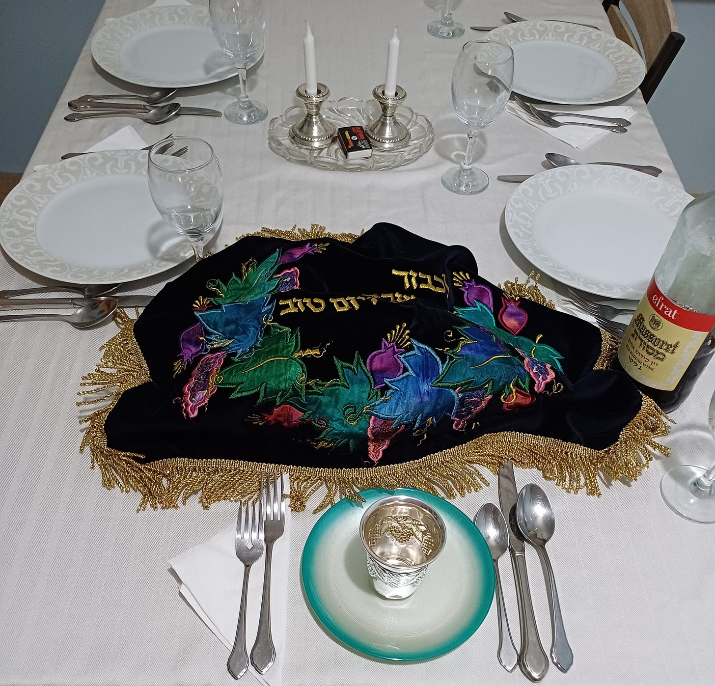 Sabbath table, china dishes on white cloth, crystal goblets, silver kiddush cup on white plate with aqua border, silver candlesticks with tall candles on glass dish, challah cover with gold fringe and lettering and appliqued grapes, vine leaves, and pomegranats in rich purples, teals and blue