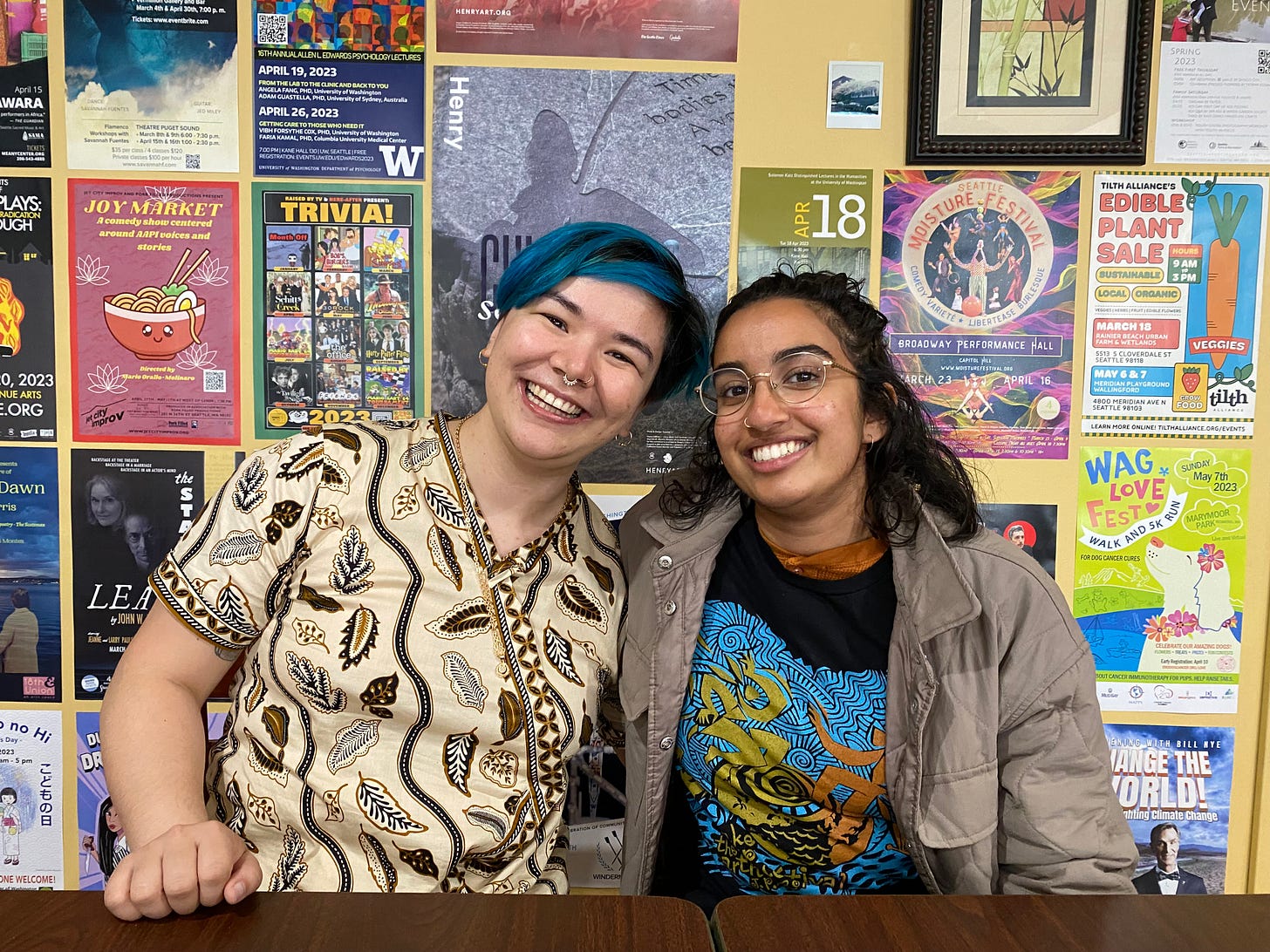 Ally and Anju smiling in front of a wall of flyers