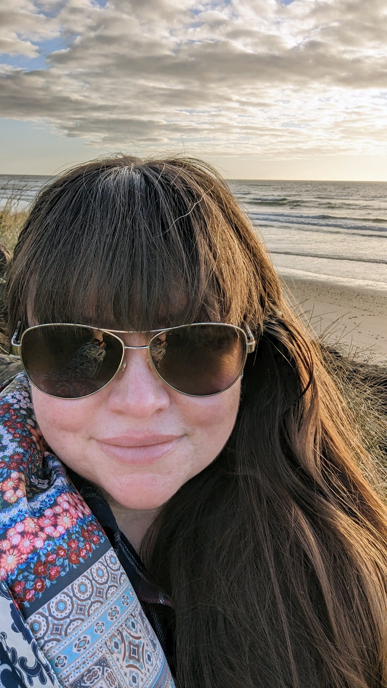 Photo of Sara, wearing sunglasses and a patterned coat, on the beach in Oregon at sunset