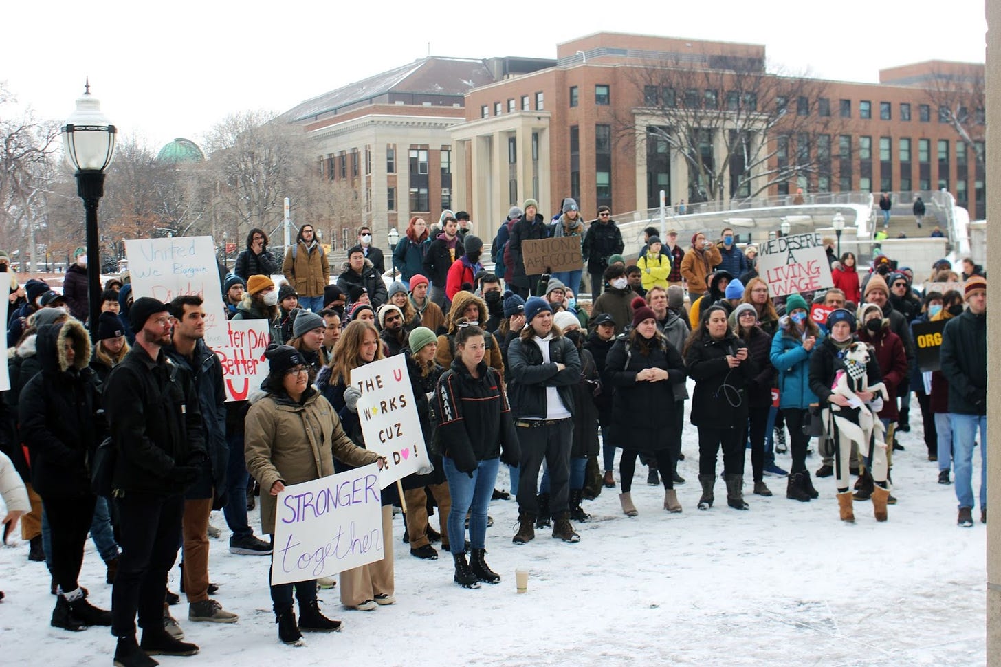 a crowd of people wearing winter garb and standing in the snow on campus in front of big brick buildings hold signs reading "we deserve a living wage," "the U works cuz we do!" and "stronger together"