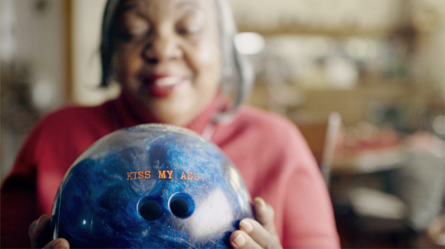 A blurred image of the protagonist Judy, a black woman with short grey hair, wearing a red sweater and holding a blue bowling ball with “Kiss My Ass” printed in gold.