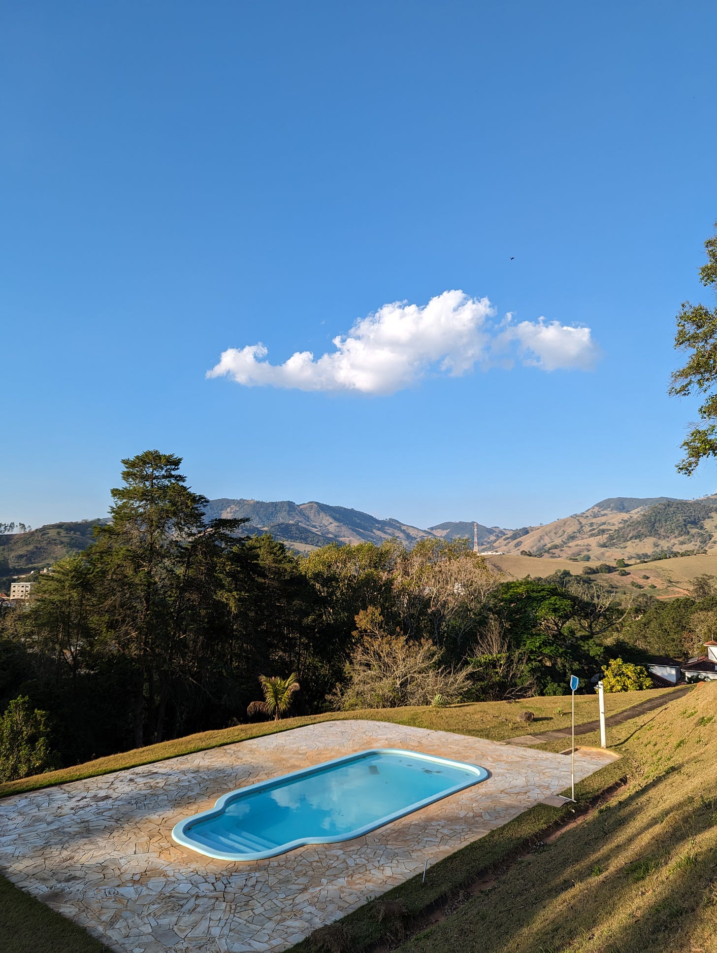 The view of a small, shallow swimming pool as seen from a hill, with trees around it and mountains in the distance. A blue sky above with just one white cloud and a black dot that appears to be a bird.