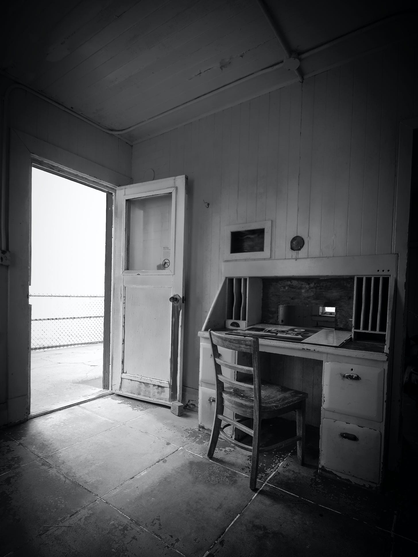 A black and white image of a vintage desk and chair in an abandoned room