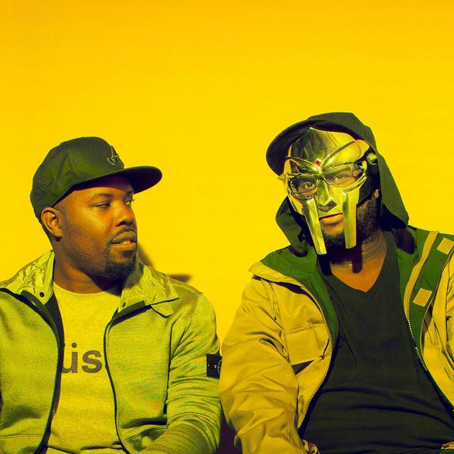 Two Black hip hop MCs seated next to each other, with a yellow background — one with baseball cap, the other with metal mask and glasses.