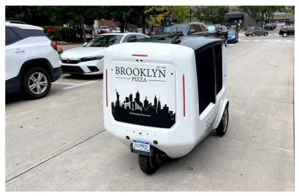 A three-wheeled white self-driving delivery vehicle. A "Brooklyn Pizza" logo is on the front