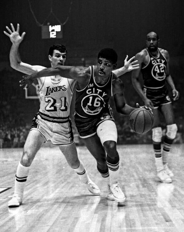 A black-and-white photo of Al Attles, wearing a dark Warriors uniform, No. 16, with an image of the Golden State Bridge on the front, dribbling the ball past a defender with his arms raised. 