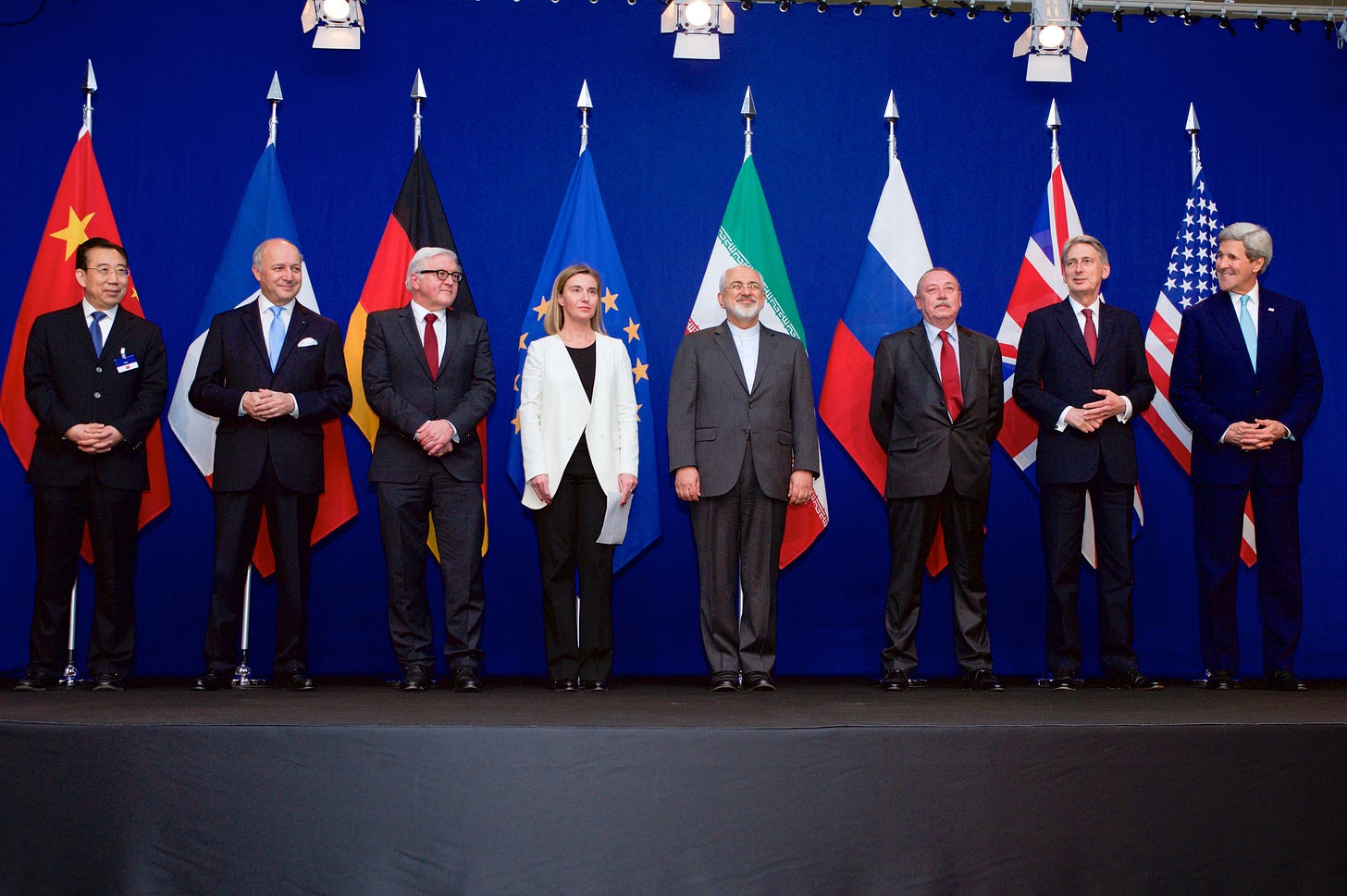 Group of officials standing on stage in front of flags from various countries, dressed in formal attire. Group of officials standing on stage in front of flags from various countries, dressed in formal attire.