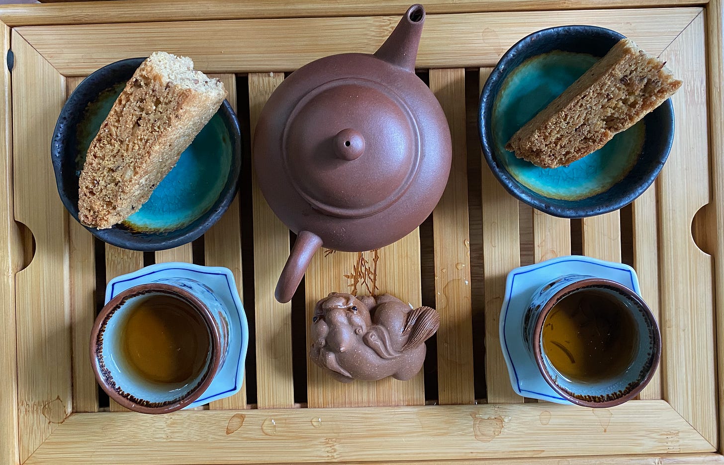 A teat tray from above: small brown teapot, brown tea dragon, two small round mugs of dark tea on saucers, and two tiny green dishes with half a biscotti in each.