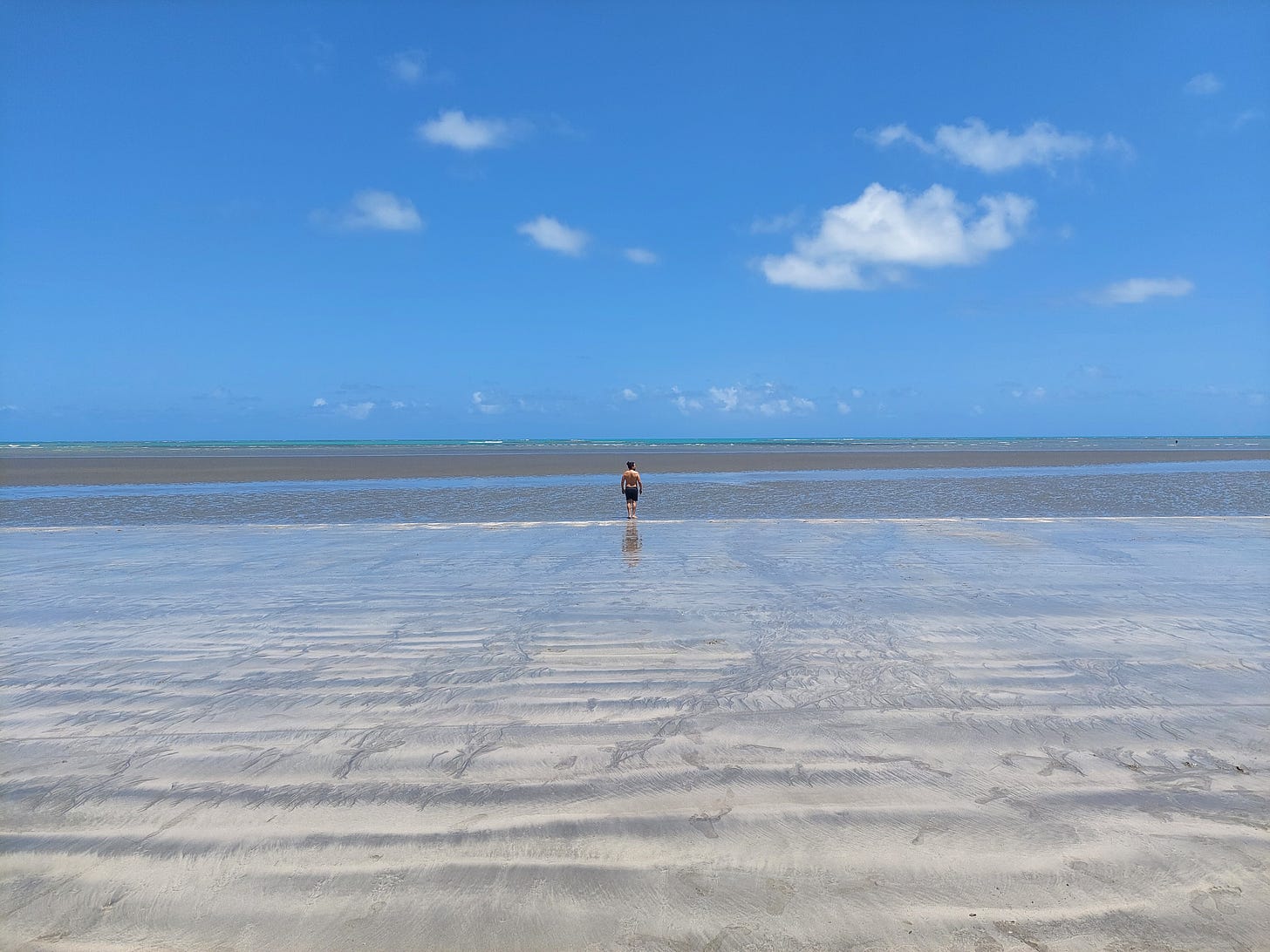 Imagem de uma praia com maré baixa e a areia molhada por quilômetros, bem rasa até o horizonte. No centro, um homem caminha de costas, distante