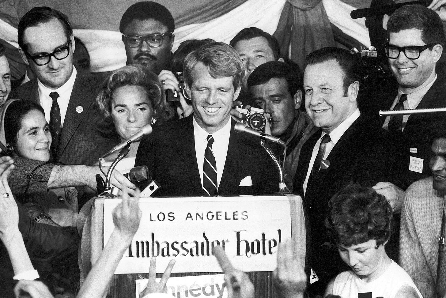 black and white photo of Rosey Grier and others standing behind Robert F. Kennedy speaking at a podium