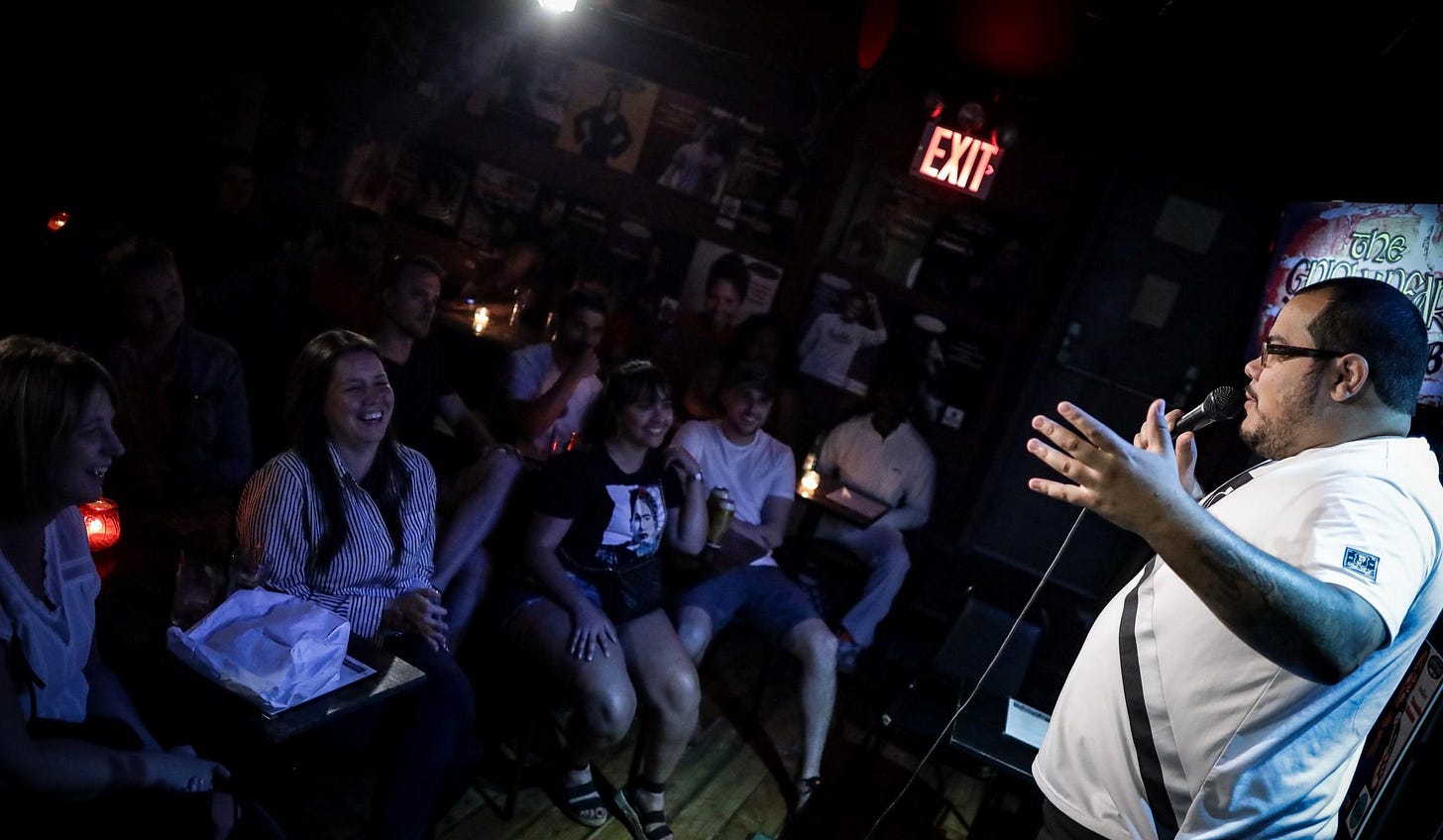 Comedian Mario Benitez on stage in a semi-dim comedy club in New York City. Benitez is a 30-something male who wears glasses. The crowd, composed of people who look his age and a bit younger, are smiling and engaged.